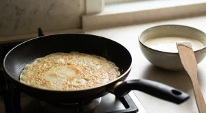 A golden crepe cooks in a black pan on a stove. Beside it is a bowl of batter with a wooden spatula resting inside