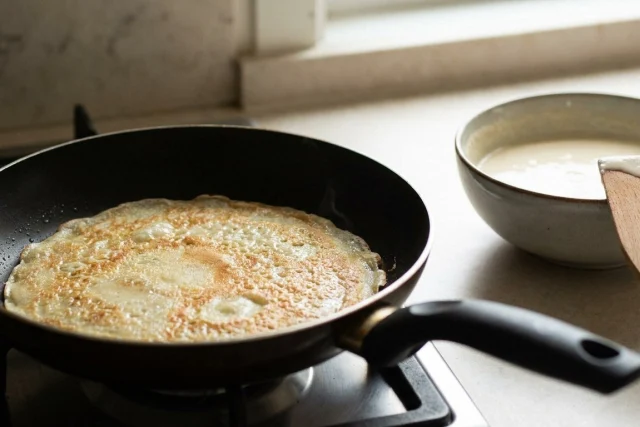 A golden crepe cooks in a black pan on a stove. Beside it is a bowl of batter with a wooden spatula resting inside